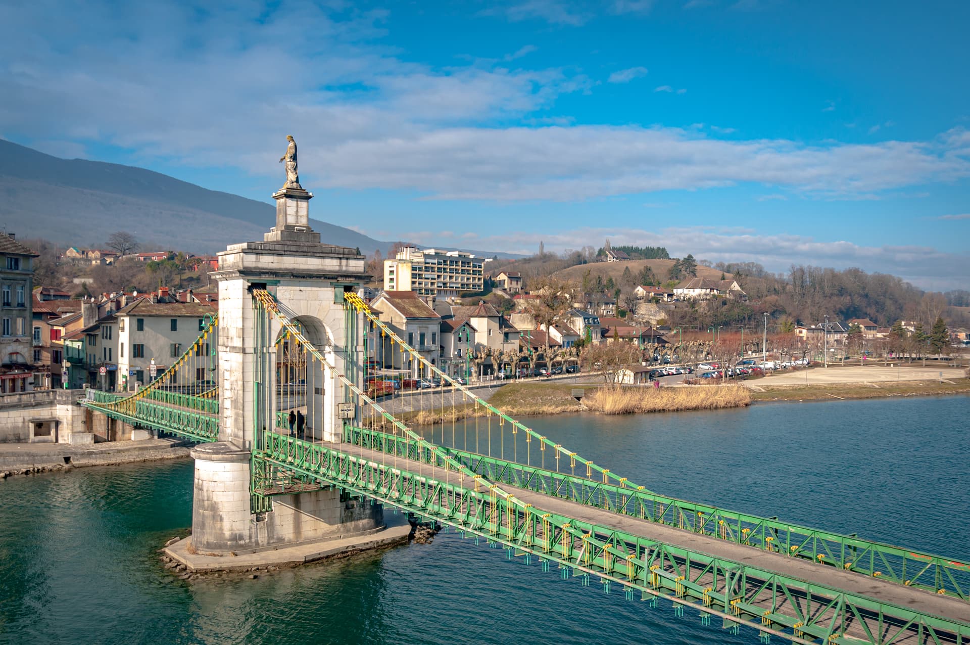 Green suspension bridge over blue water with a town and hills in the background, Seyssel.