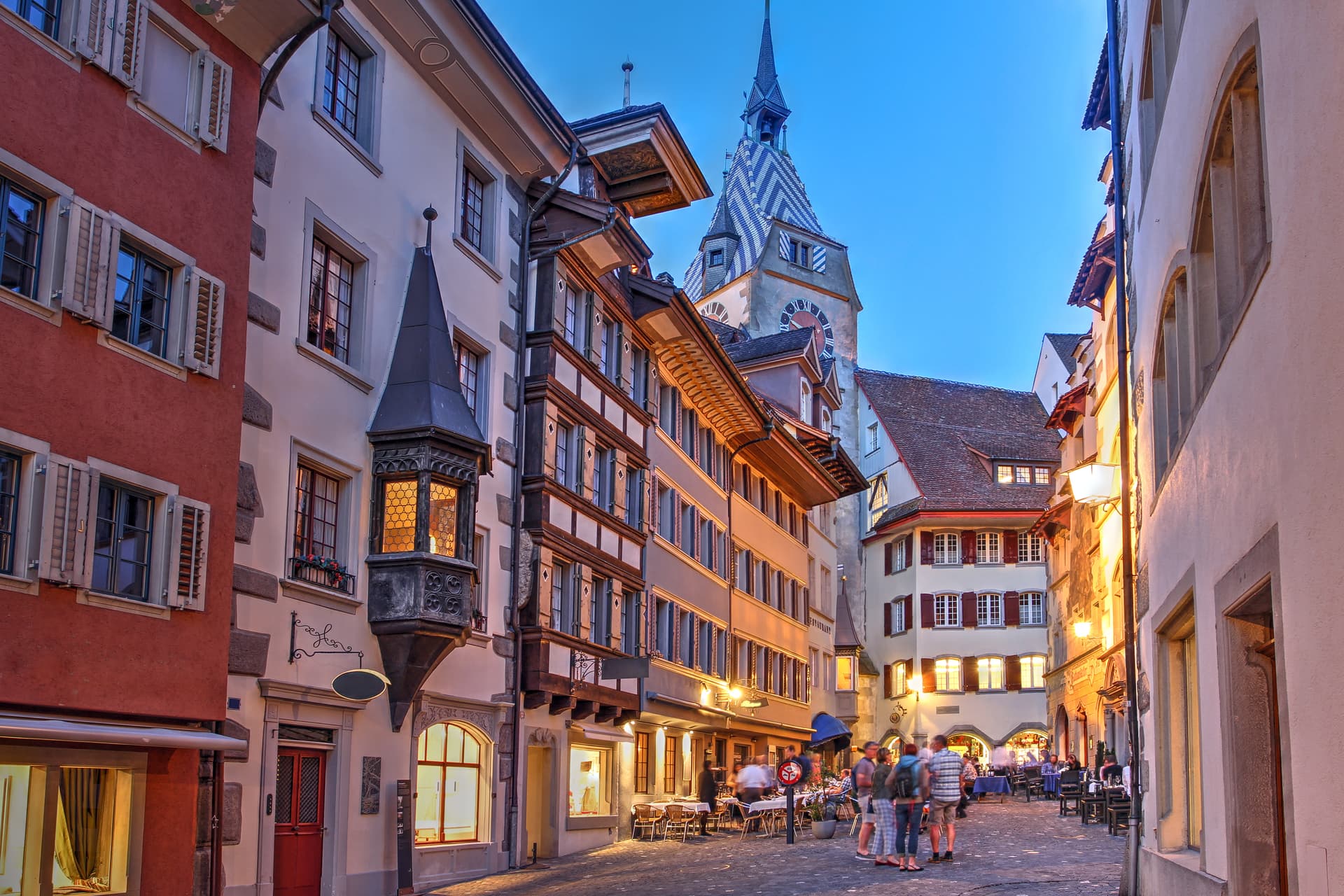 Cobblestone street in 5-Zug old town, Switzerland, with historic buildings and outdoor dining at dusk.