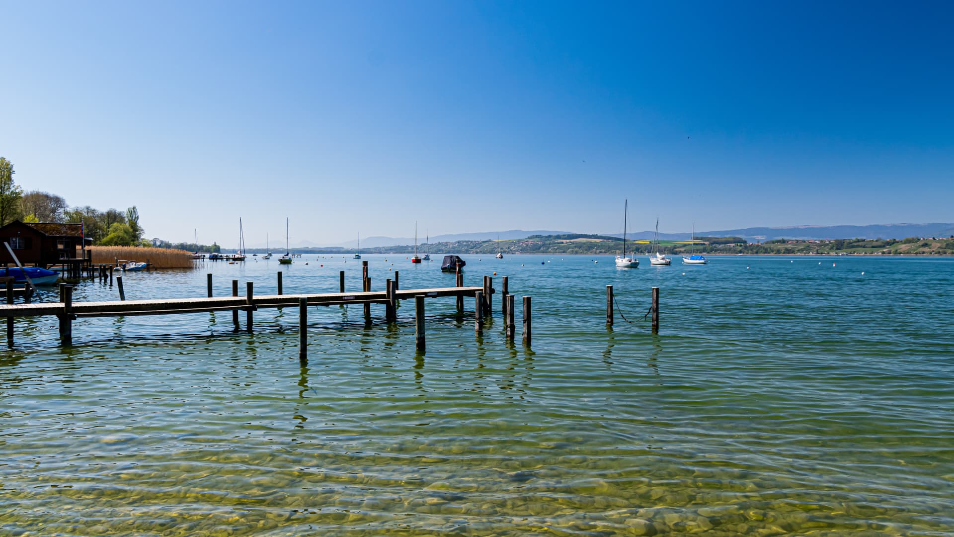 Wooden dock extending into clear lake water with moored sailboats and distant hills, Lake Murten.