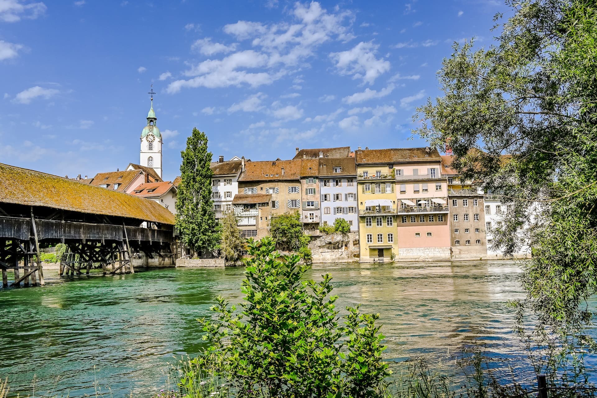 Olten covered wooden bridge, colorful buildings, and church tower over the Aare River in Switzerland.
