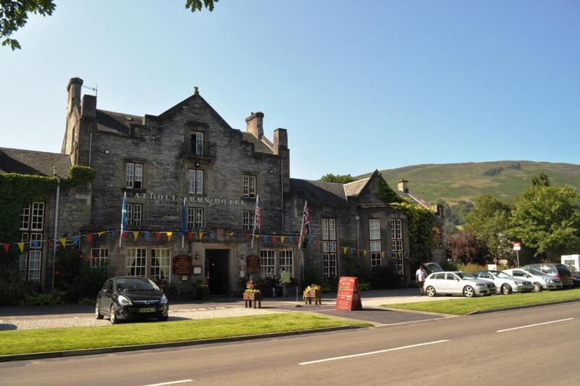 Atholl Arms Hotel stone building with flags and parked cars against a green hillside.