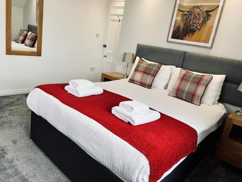 Hotel room with double bed, red throw, plaid pillows, and Highland cow artwork.