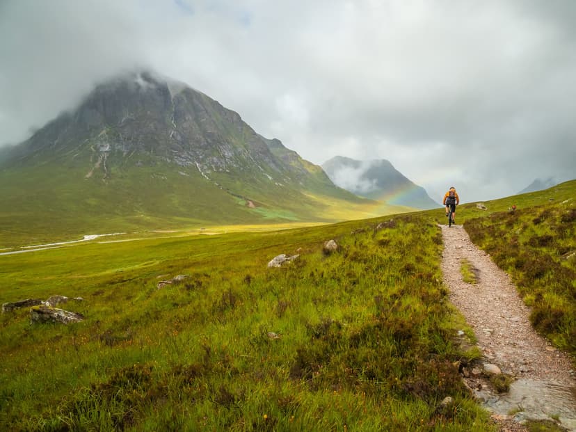 Mountain biker cycling on dirt path through green valley with mountains and rainbow in Glencoe.