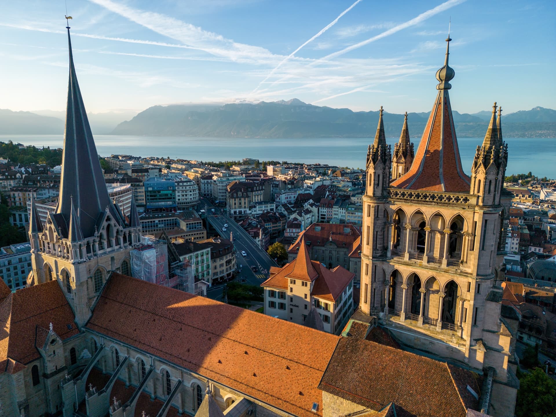 Lausanne Cathedral towers overlooking Lake Geneva and distant mountains under a blue sky.