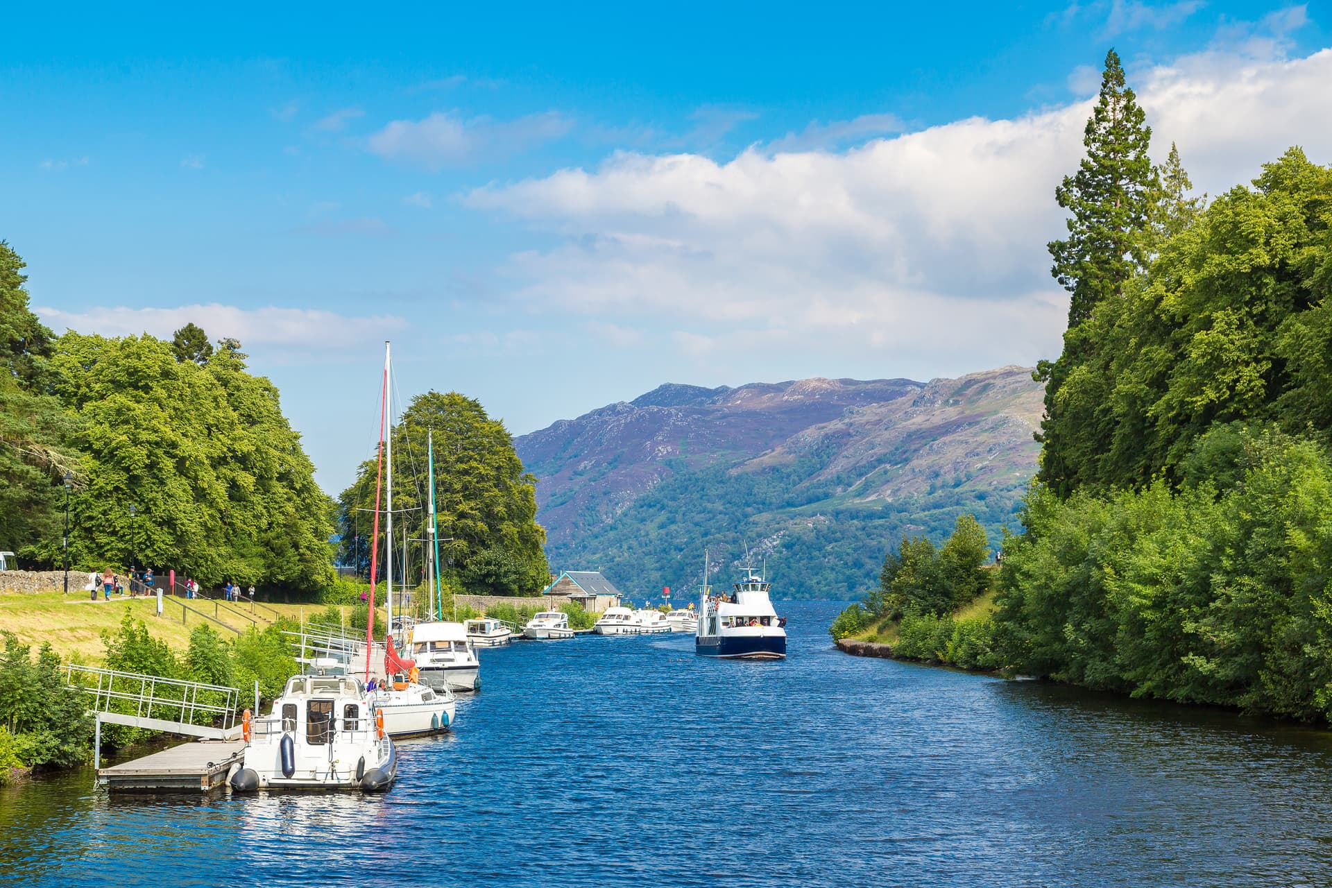 Boats docked and cruising on water channel near Loch Ness with green trees and mountains.