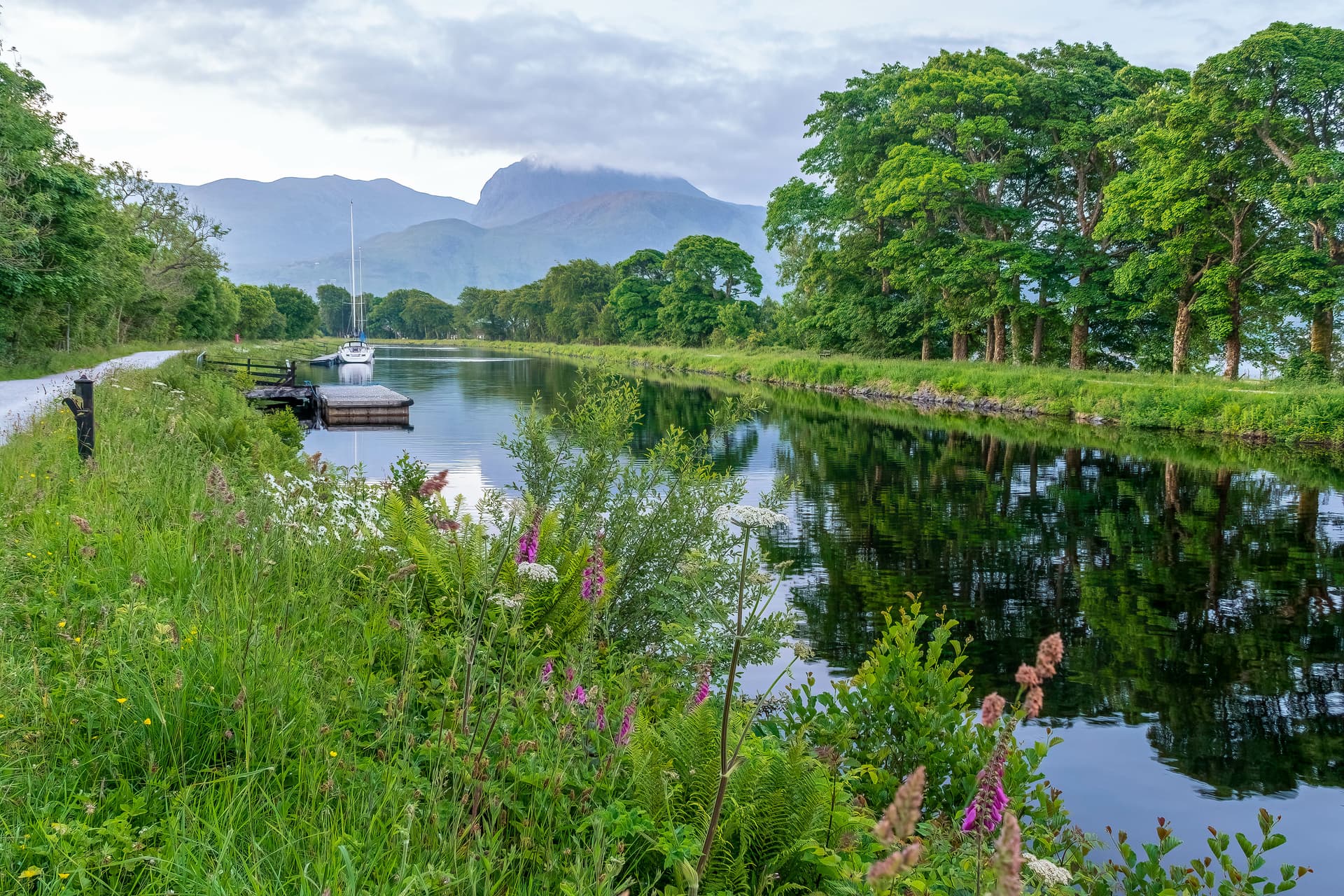 Caledonian Canal bike path with sailboat docked, lush greenery, and misty mountains in background.