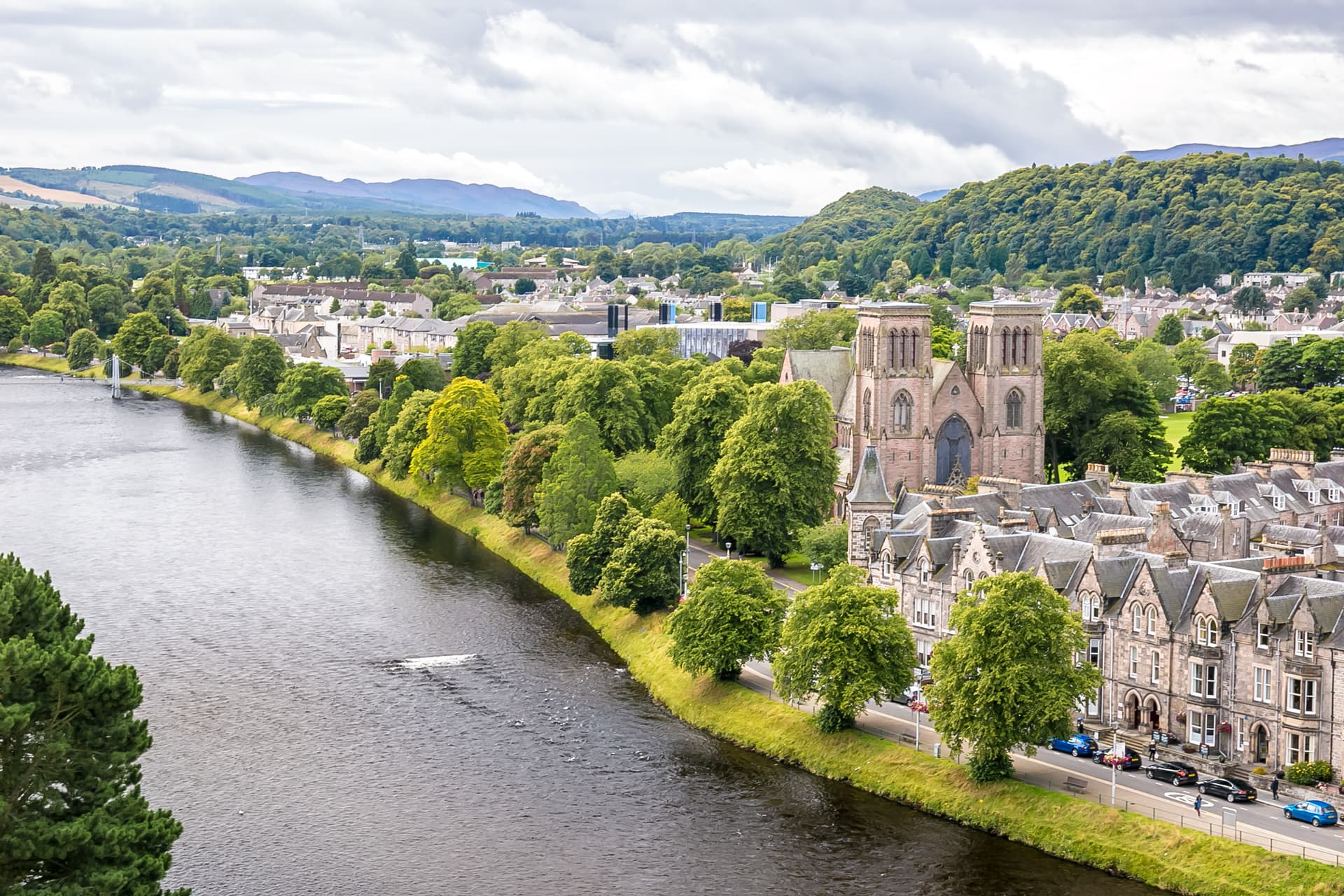 River view of Inverness with stone church, green trees, and distant hills under cloudy sky.