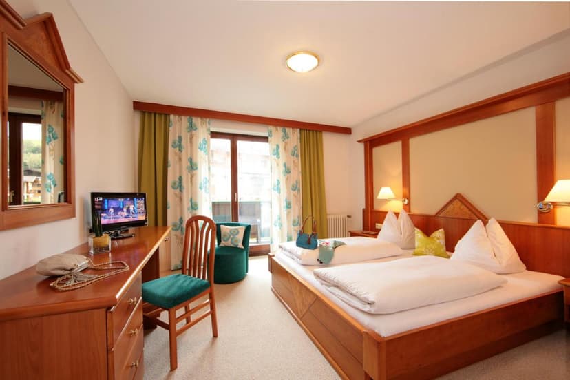 Hotel room with wooden furniture, twin beds, TV, and balcony access in Kaprun.