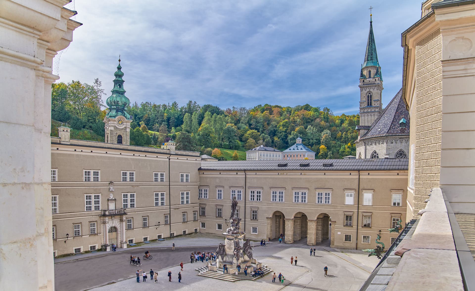 Historic square in Salzburg with buildings, monument, and forested hill under a blue sky.