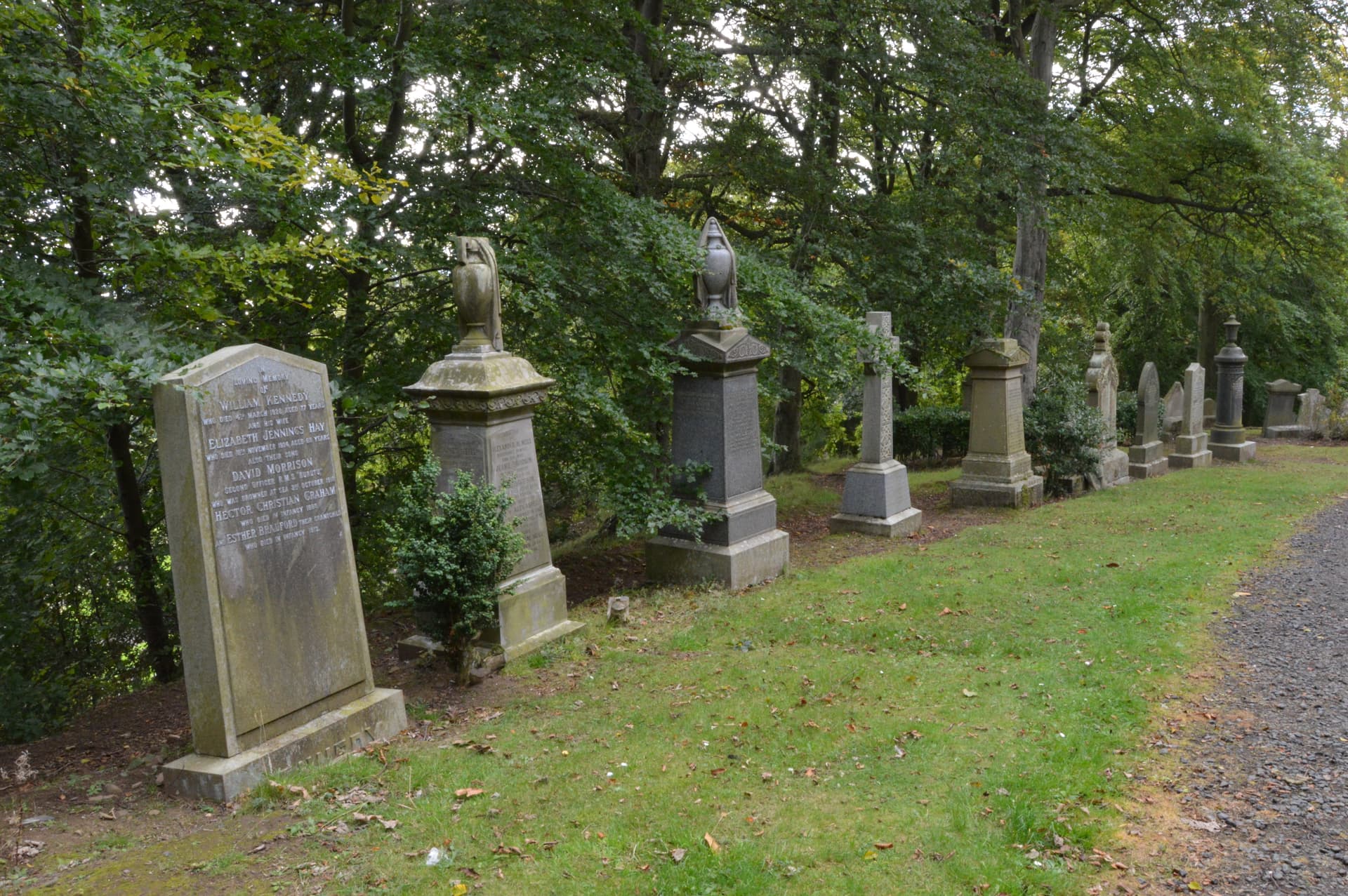 Gravestones and monuments line a grassy path under dense green trees at Balgay Cemetery, Dundee.