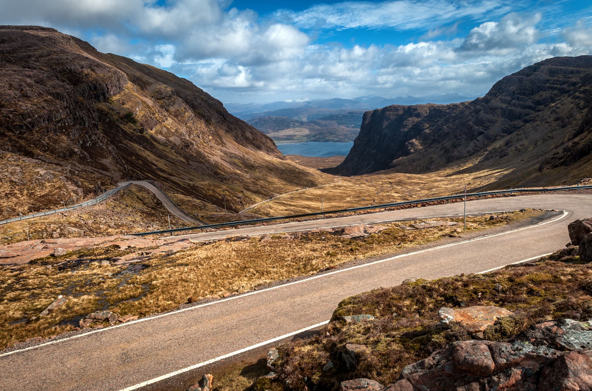 Winding mountain road descent with guardrails overlooking a valley and distant loch under a cloudy blue sky.