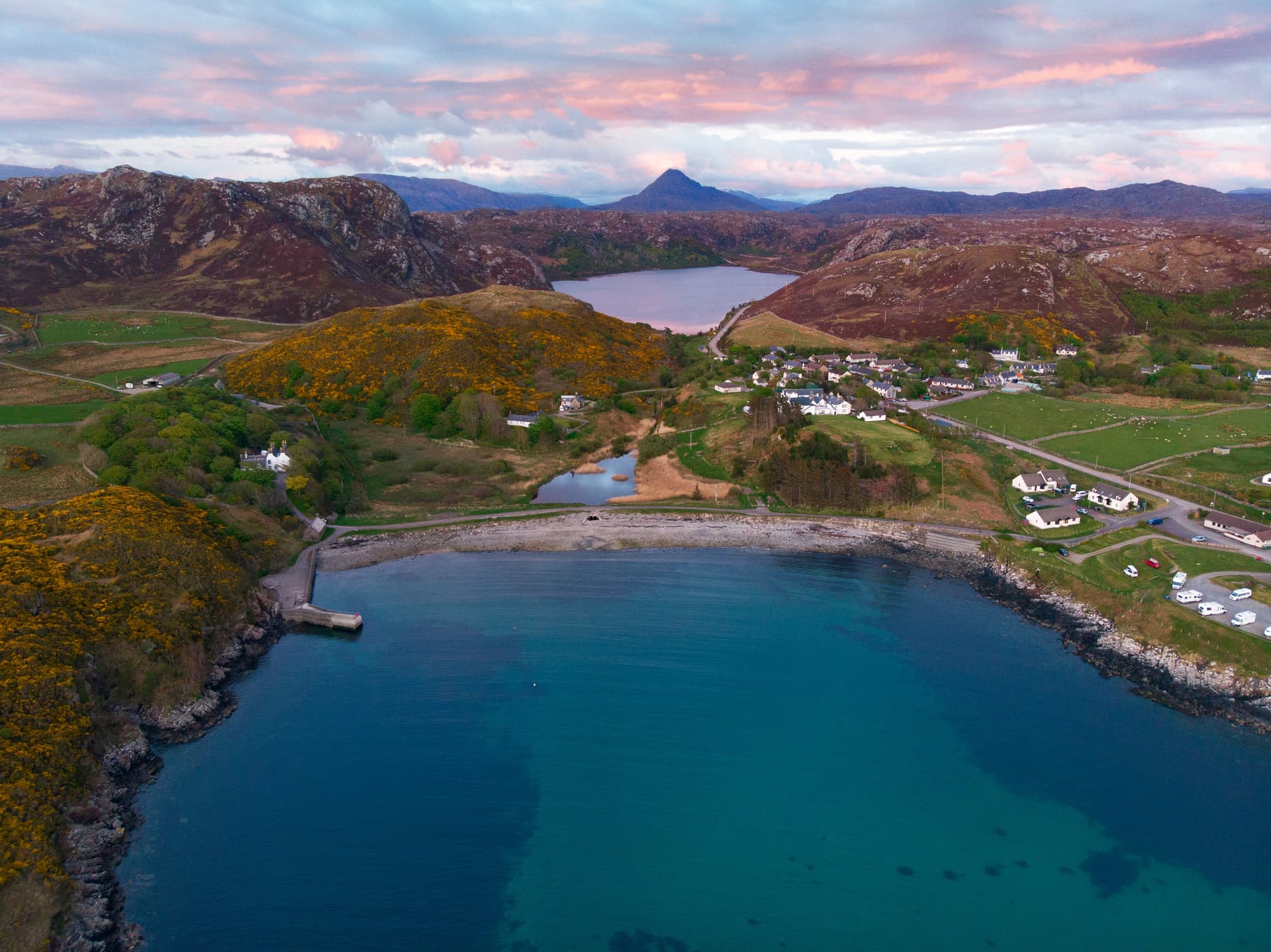Aerial view of Scourie Bay with turquoise water, coastal village, and rugged hills under a pink-tinged sky.