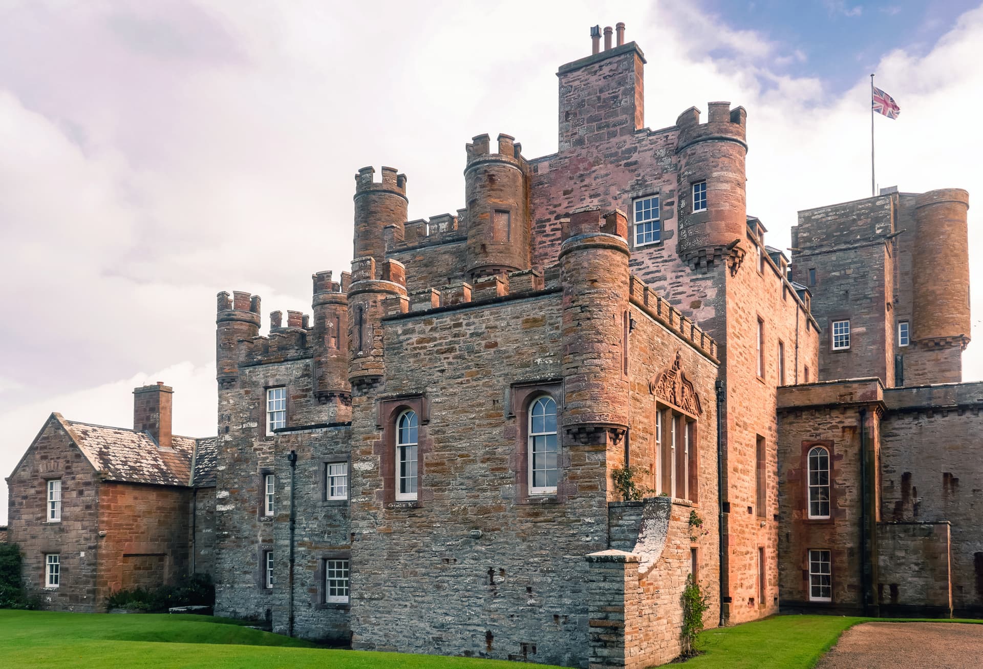 Mey Castle with turrets and stone walls, flying the Union Jack flag under a cloudy sky.