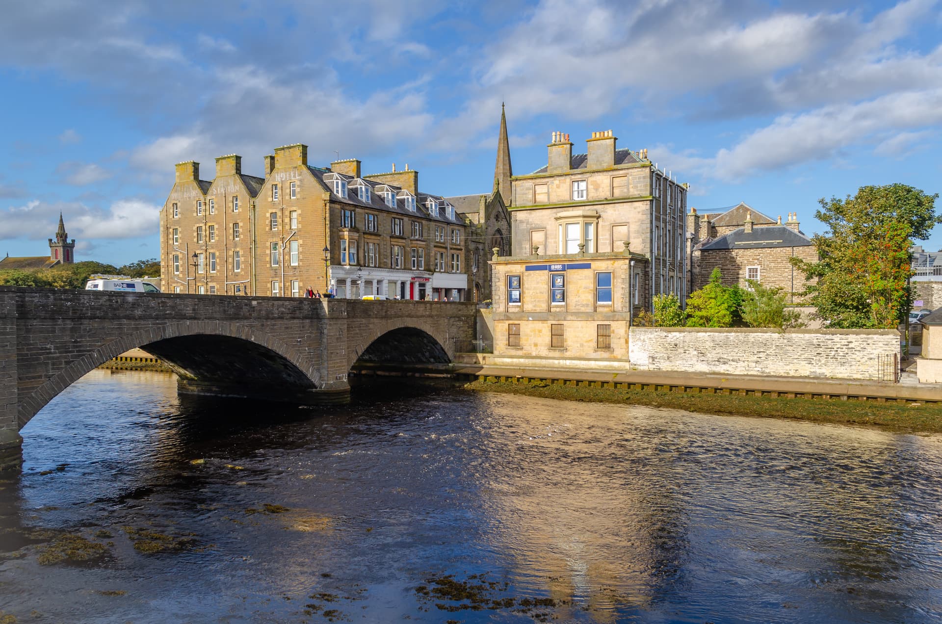 Stone bridge over river next to historic buildings and church spire in Wick, Scotland.