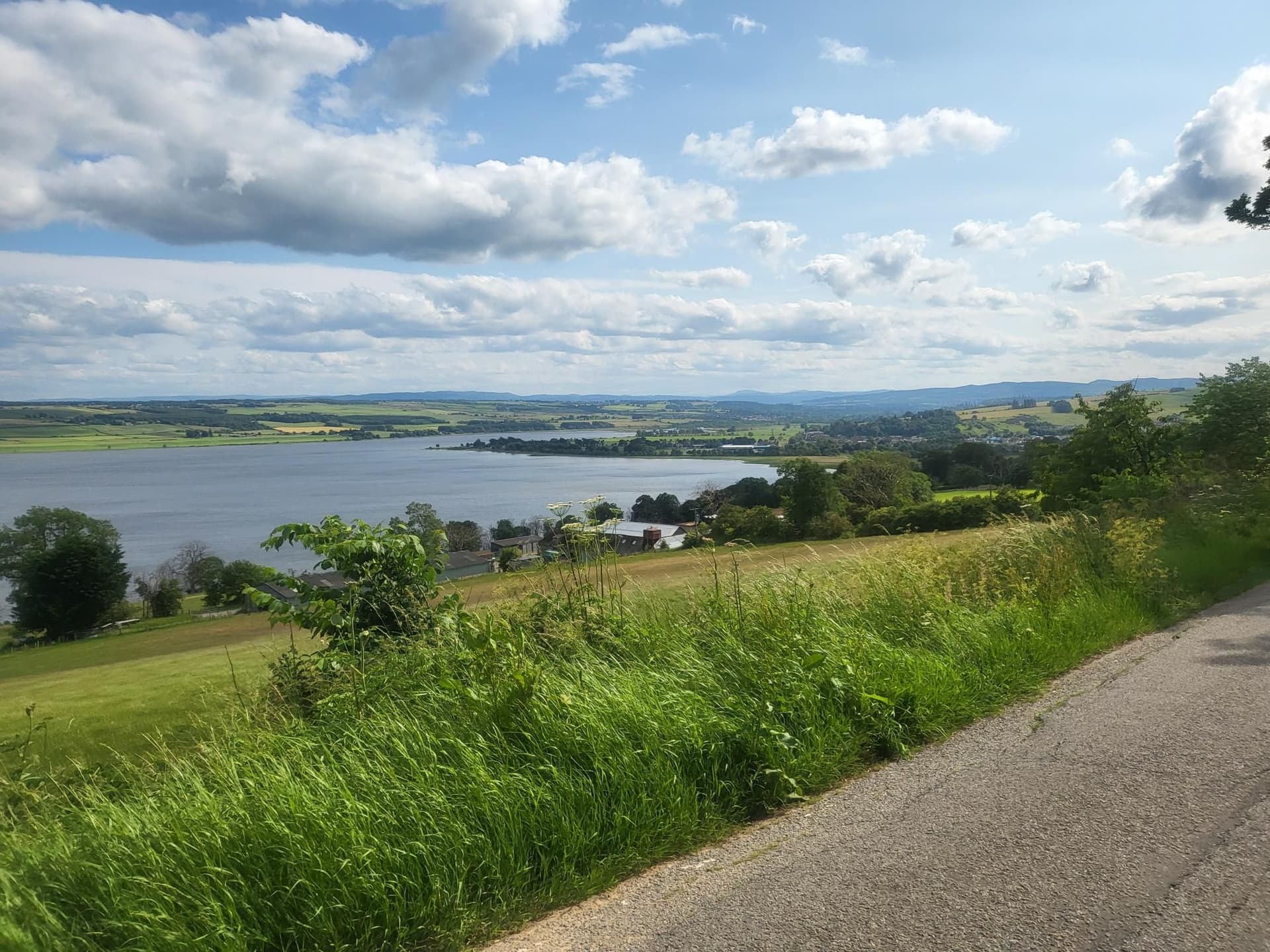 Roadside view of a large loch or lake with rolling green hills and scattered buildings under a cloudy sky.