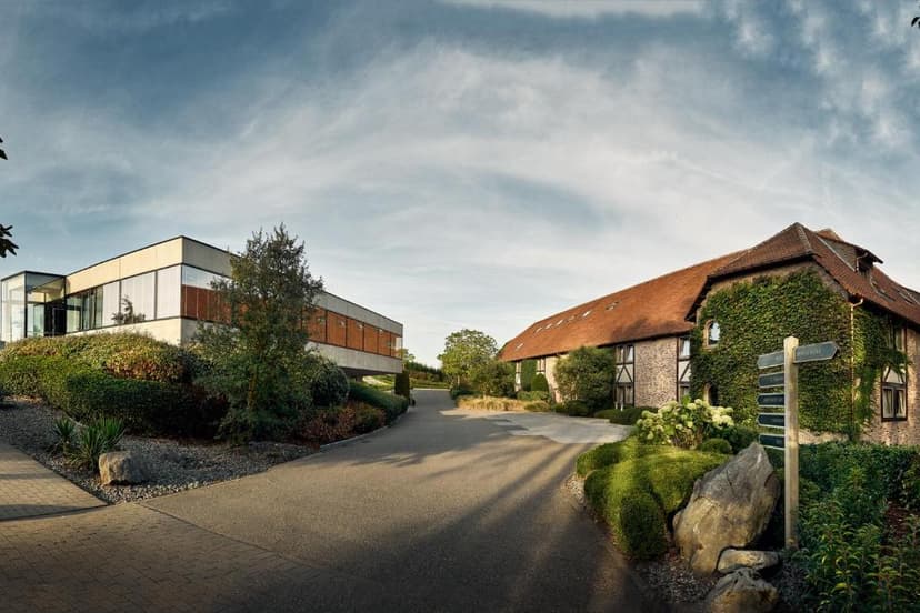 Driveway leading to modern and traditional buildings with lush landscaping under a cloudy sky.