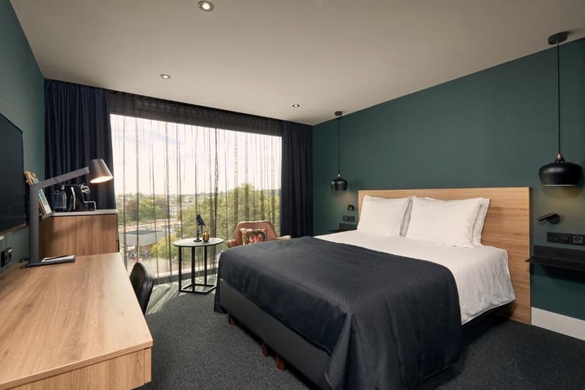 Modern hotel bedroom with large window view, dark teal walls, wood desk, and black bedding.
