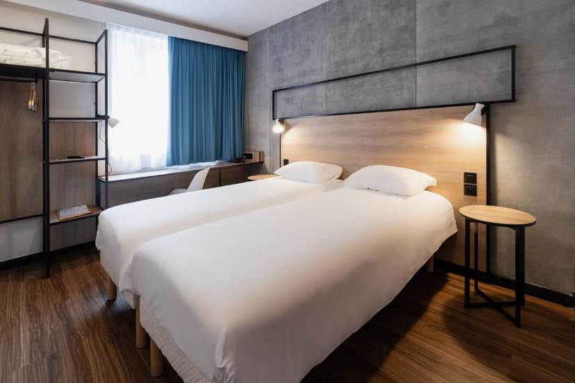 Twin beds with white linens in a modern hotel room with wood floors and blue curtains.