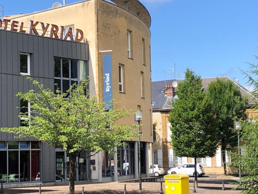 Hotel Kyriad building exterior with green trees and a yellow post box on a sunny day.