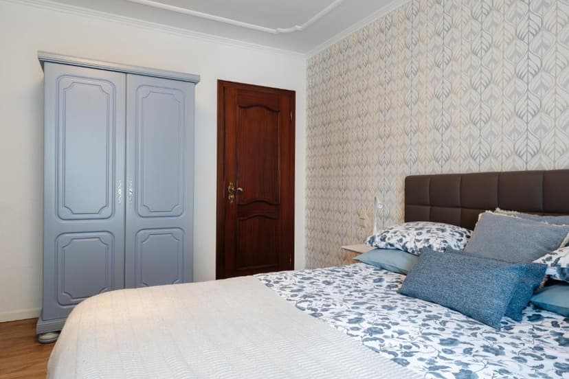 Bedroom with blue floral bedding, light blue wardrobe, and leaf-patterned wallpaper.
