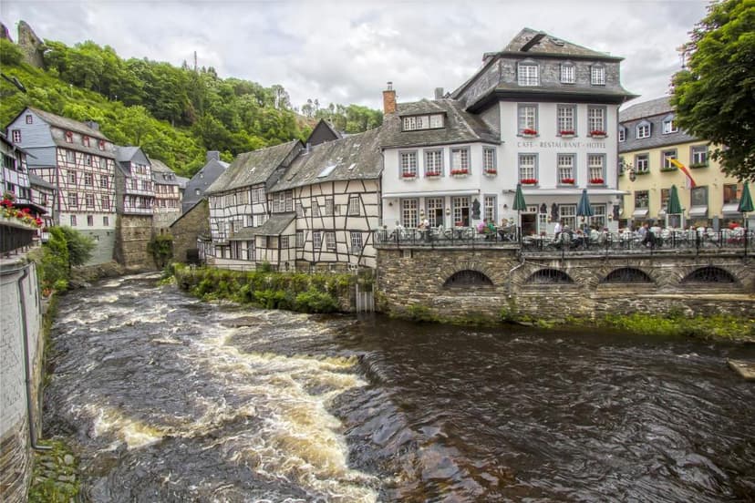 Half-timbered buildings line a fast-flowing river in Monschau with an outdoor cafe terrace.