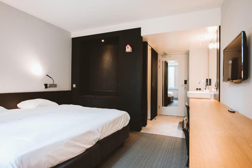 Modern hotel room with white bedding, dark accents, wall-mounted TV, and open vanity area