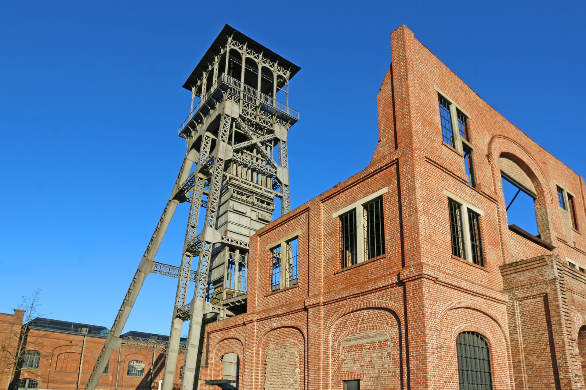 Industrial mine headframe tower next to partially ruined red brick industrial building under clear blue sky.