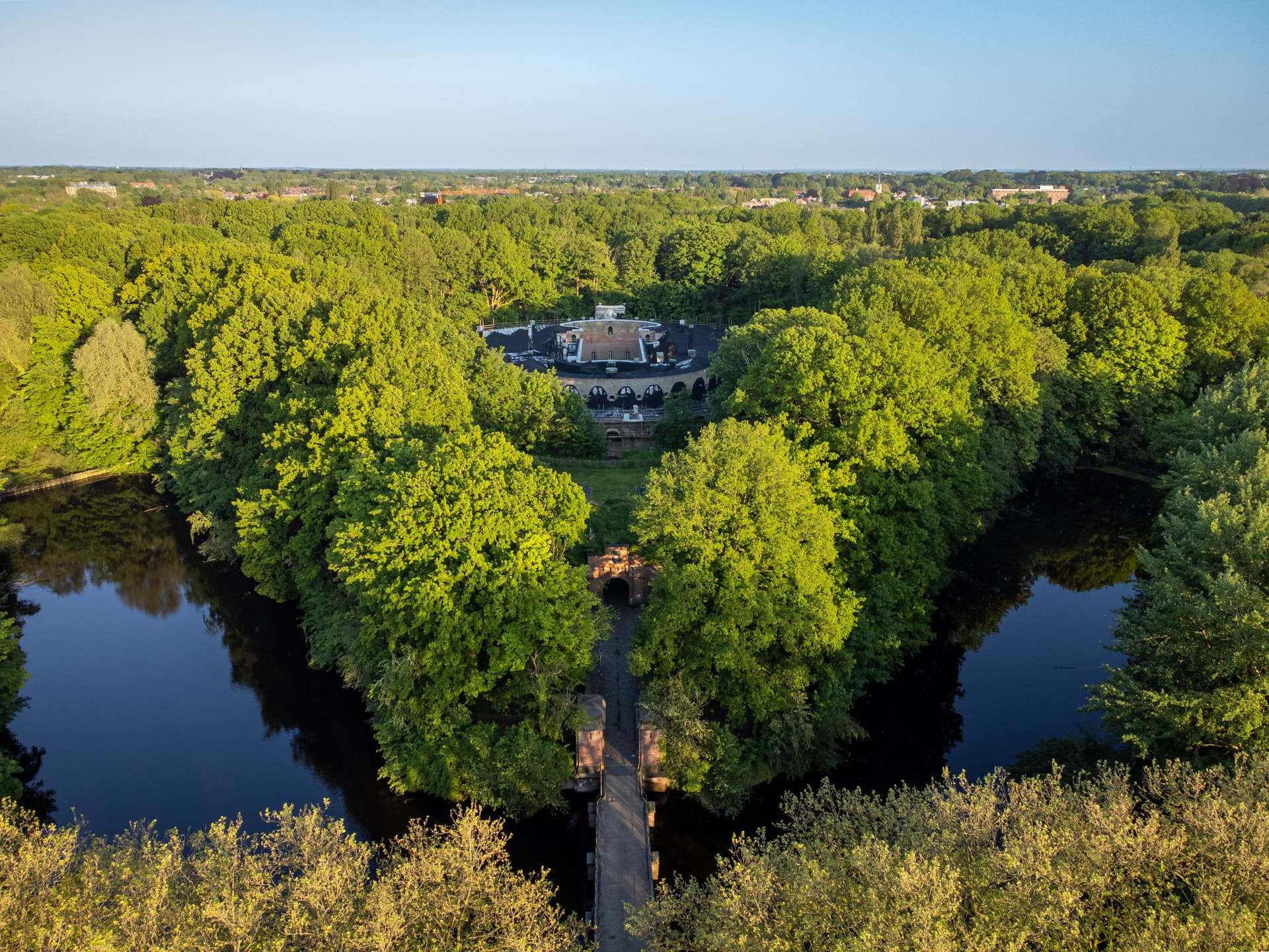 Historic building surrounded by dense green trees and dark water moat, aerial view.