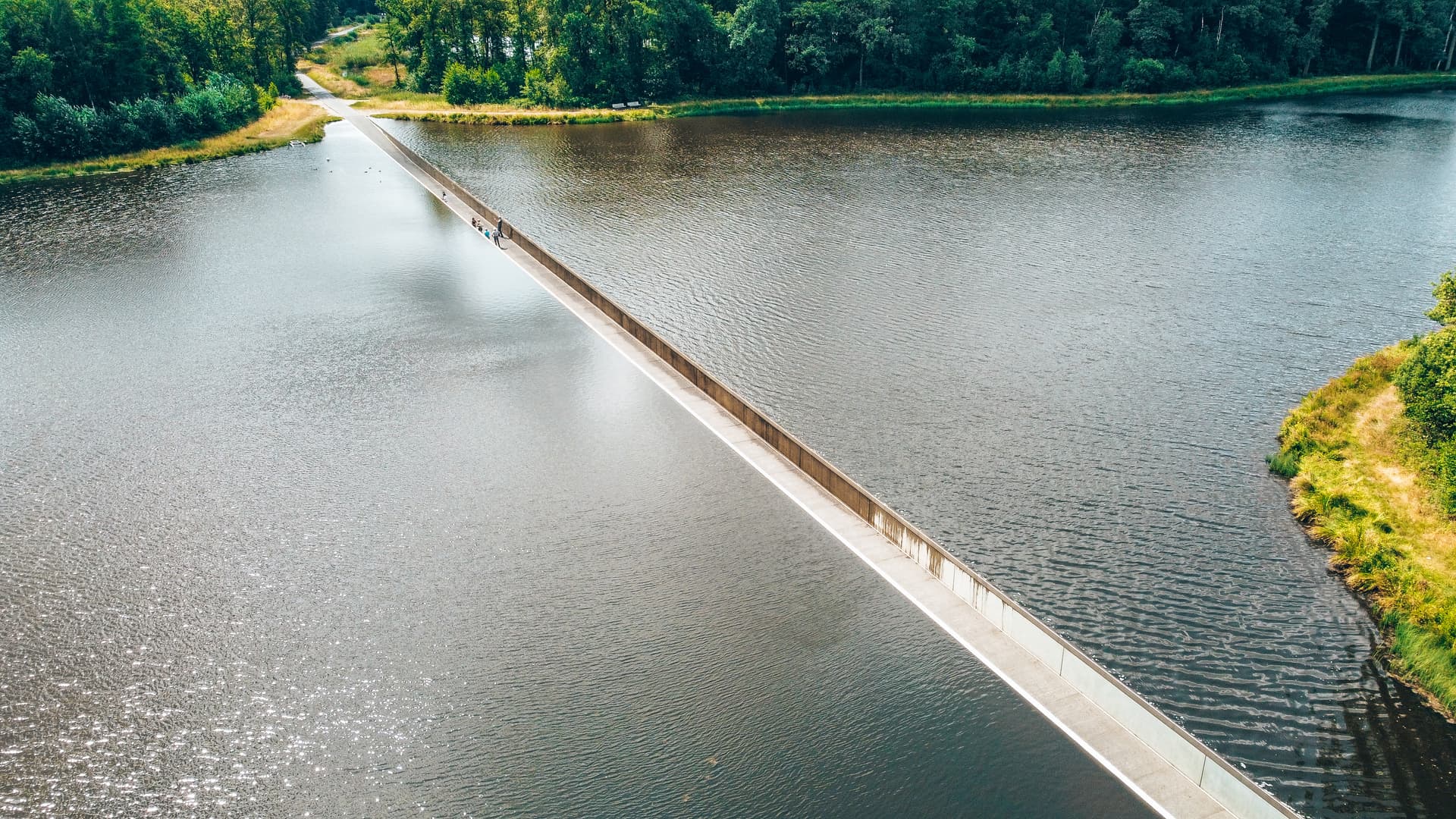 People walking along a concrete dam separating two bodies of water surrounded by green forest.