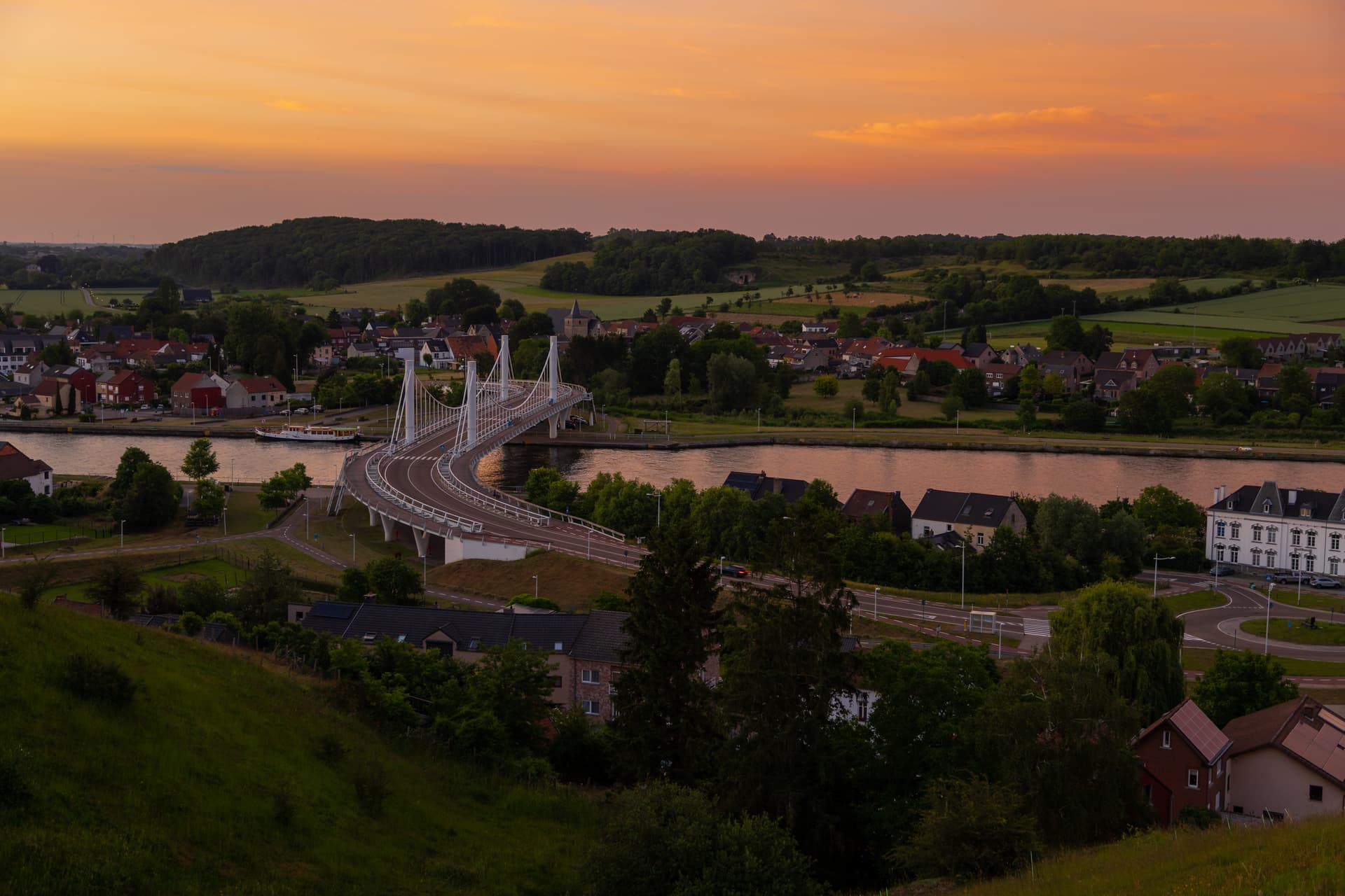 Modern cable-stayed bridge over river near town at sunset with orange sky