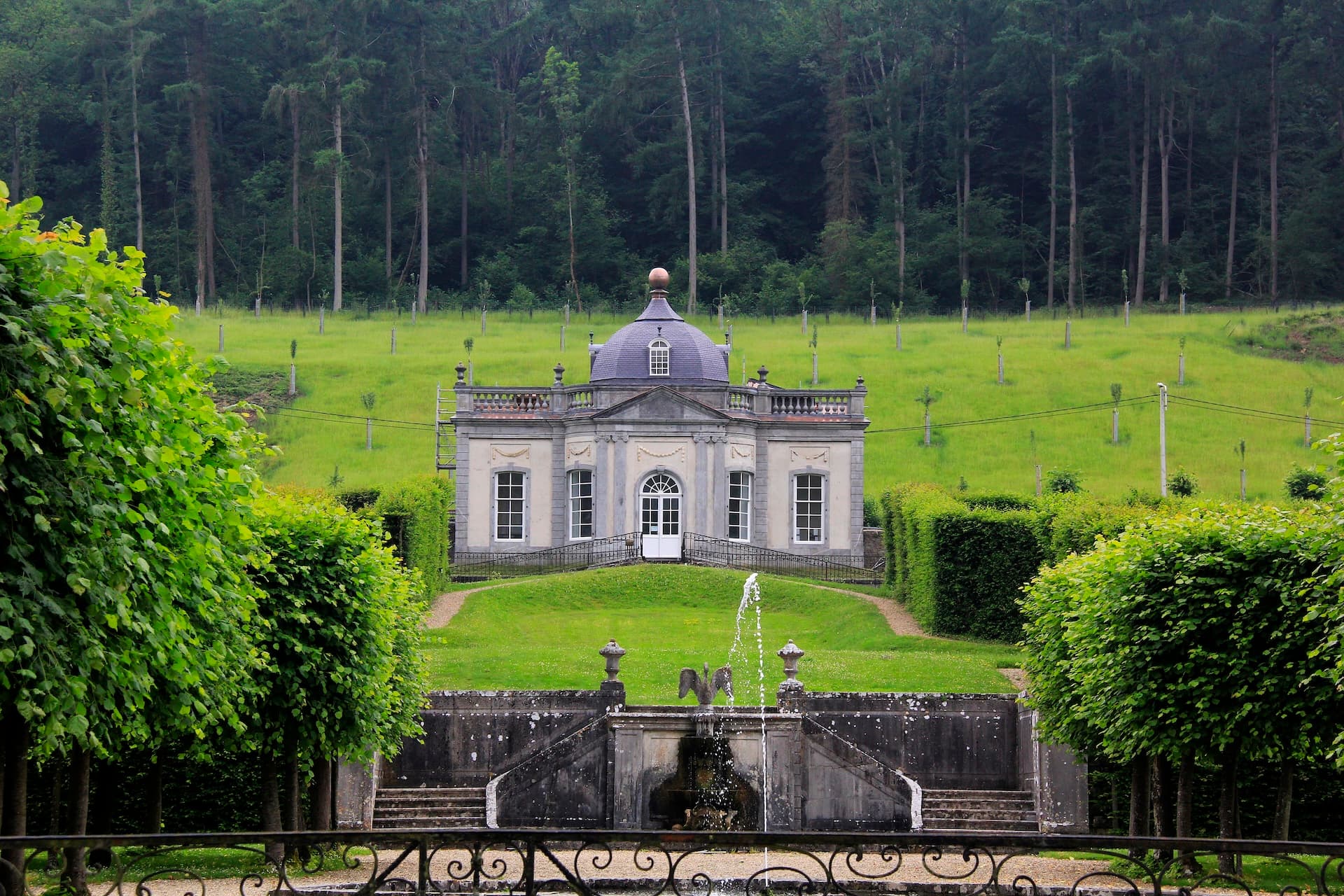 Chateau Freyr pavilion with fountain, manicured hedges, and dense forest backdrop.