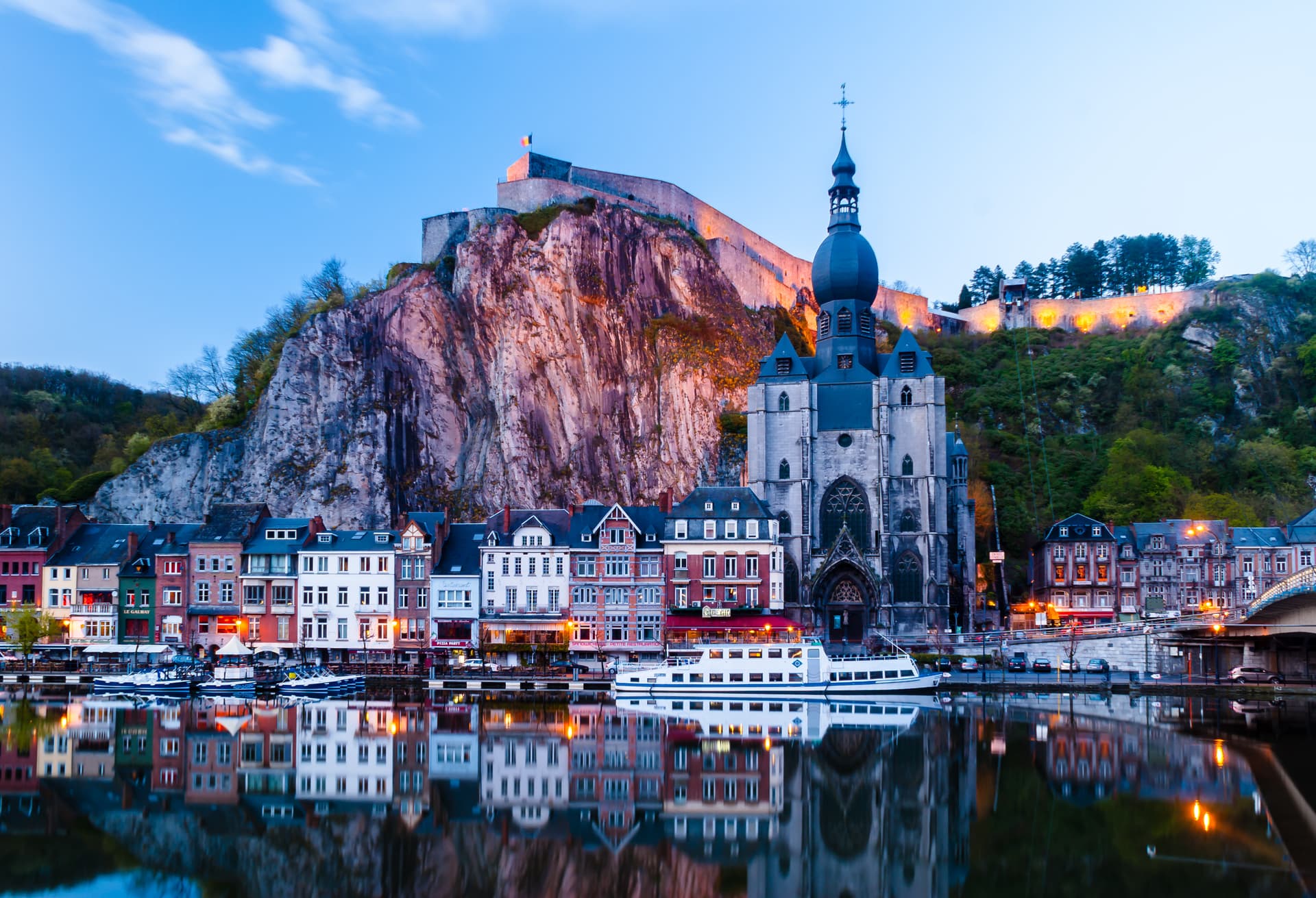 Town with illuminated citadel and church reflected in river at twilight, Dinant.