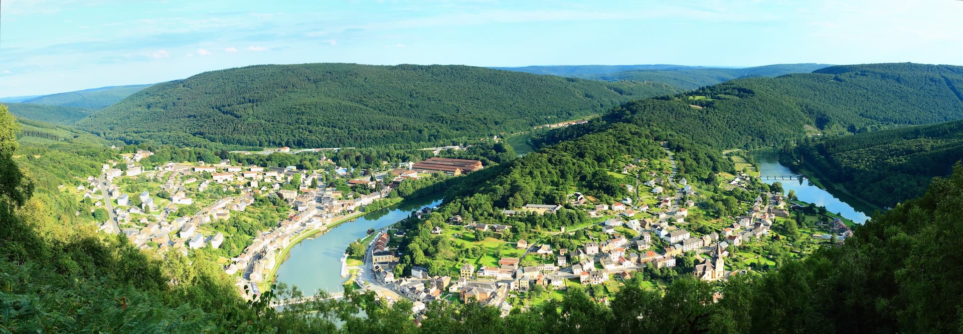 Town nestled in a river valley surrounded by dense green forested hills under a blue sky.