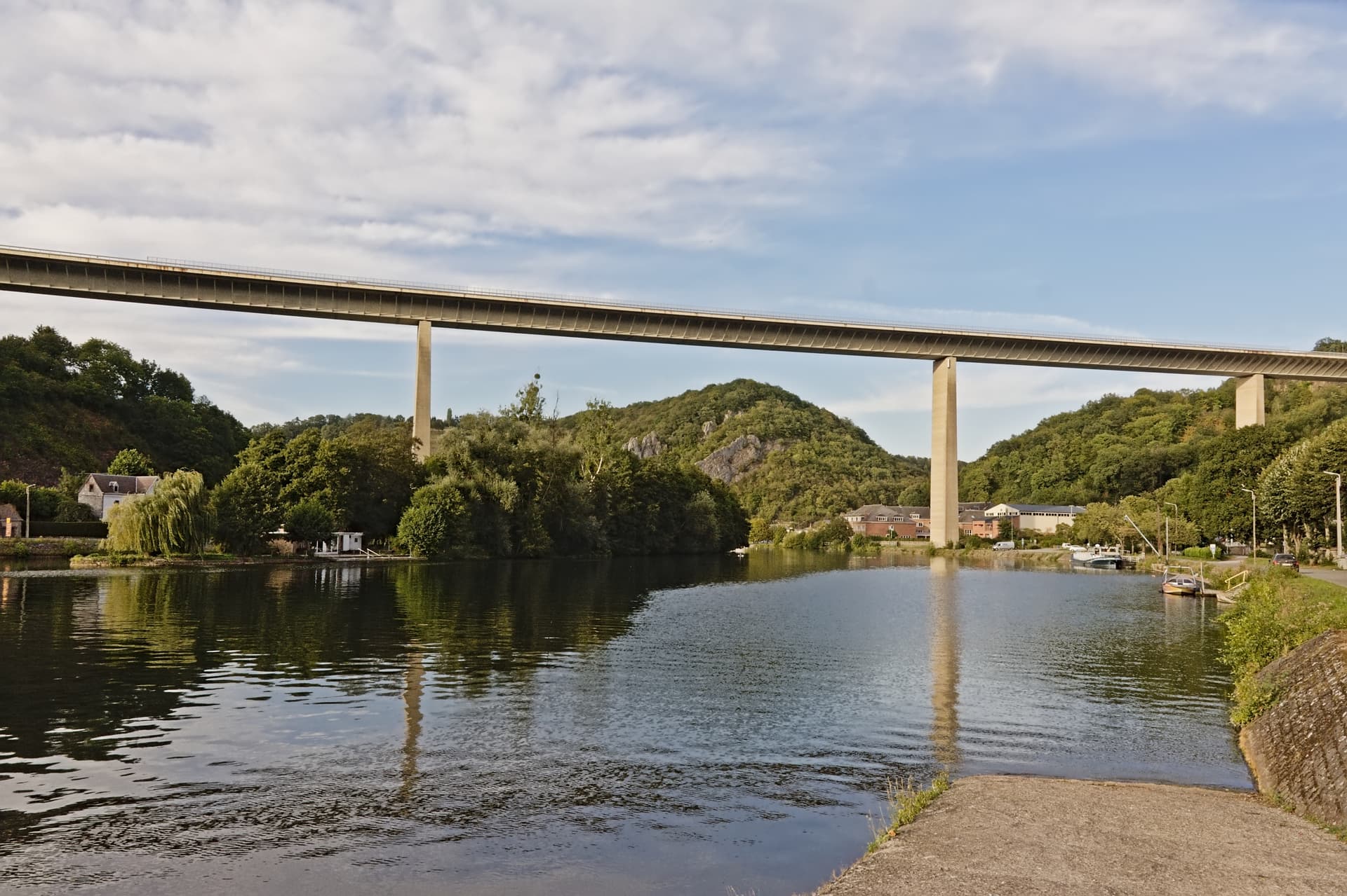 Viaduct bridge spanning a river valley with lush green hillsides and small boats docked.