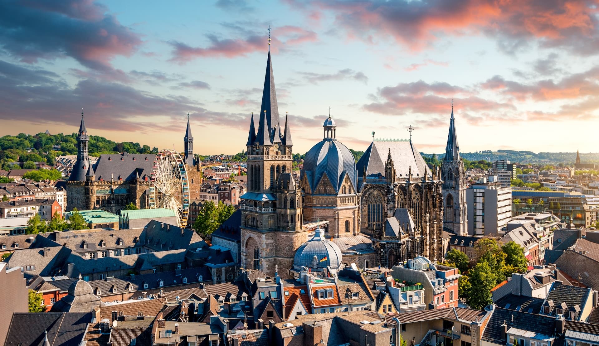 Aachen Cathedral and city rooftops with a Ferris wheel under a dramatic sunset sky.