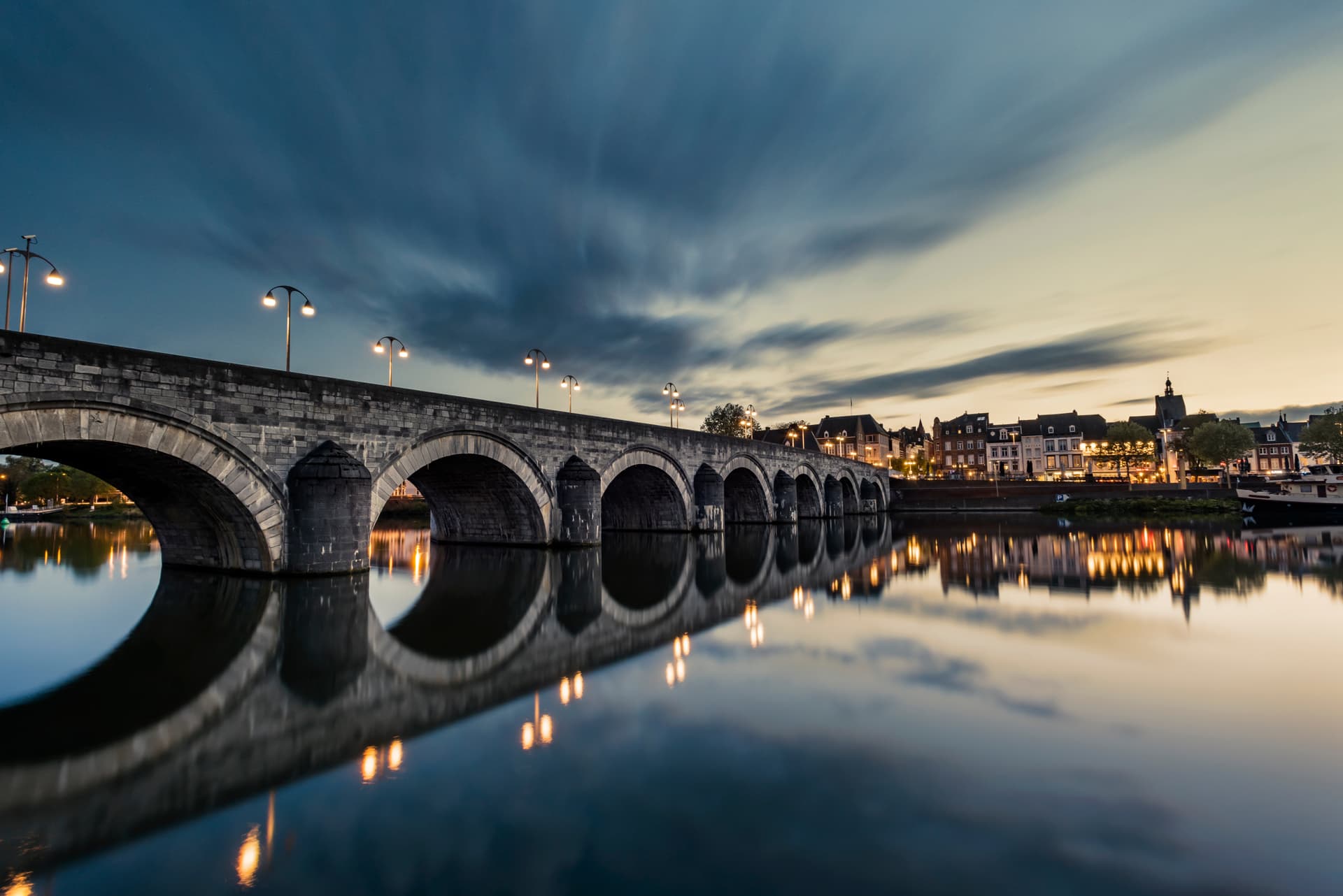Stone arched bridge illuminated at dusk reflecting in calm water near Maastricht buildings.