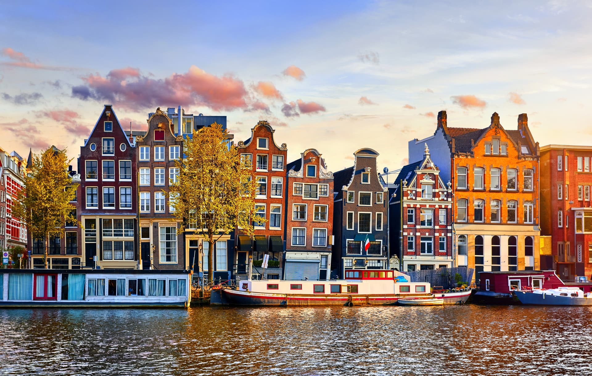 Canal houses and houseboats along the water in Amsterdam at sunset with yellow autumn tree.