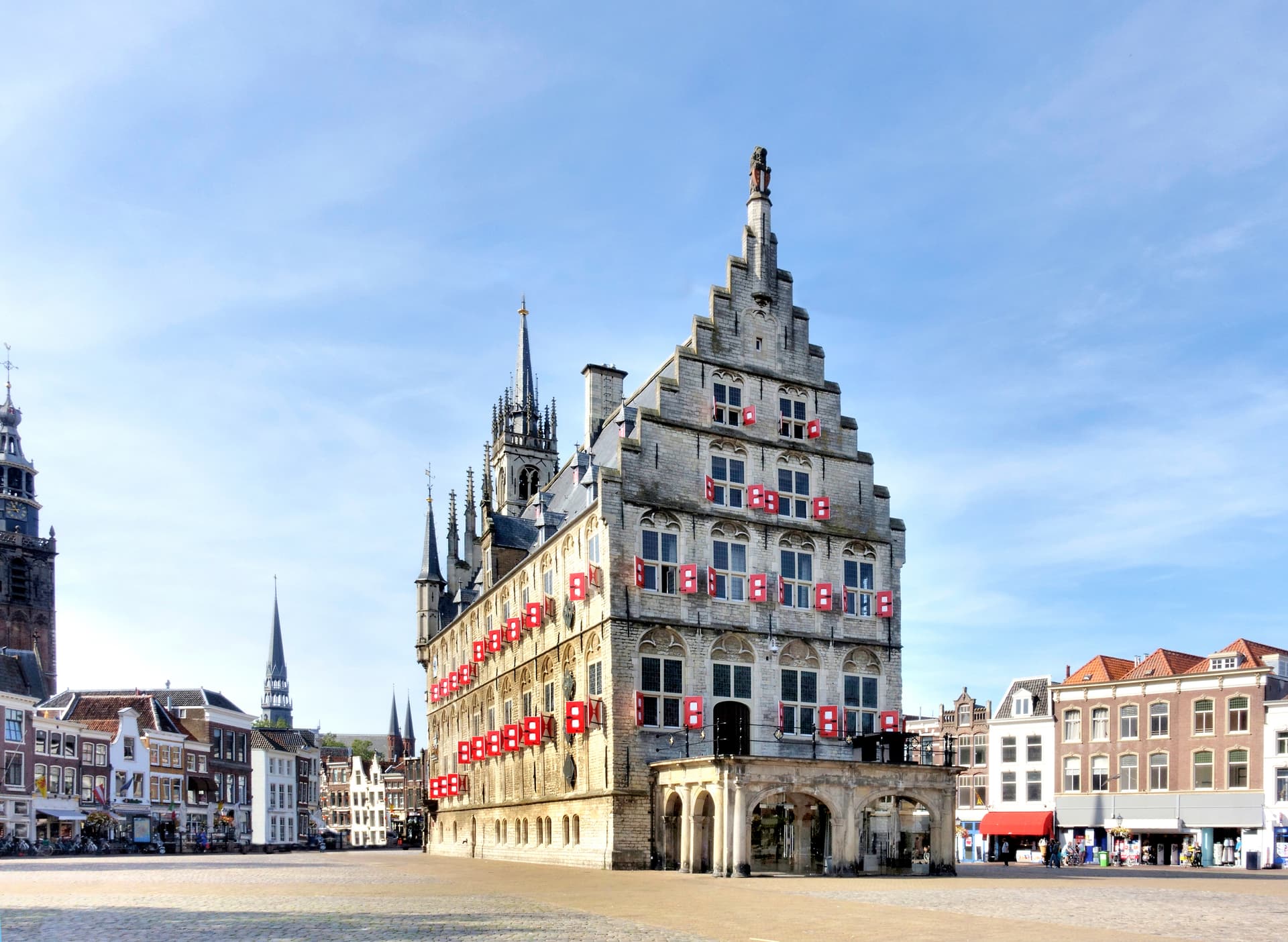 Gothic town hall with red shutters on a square, with church spires in the background.