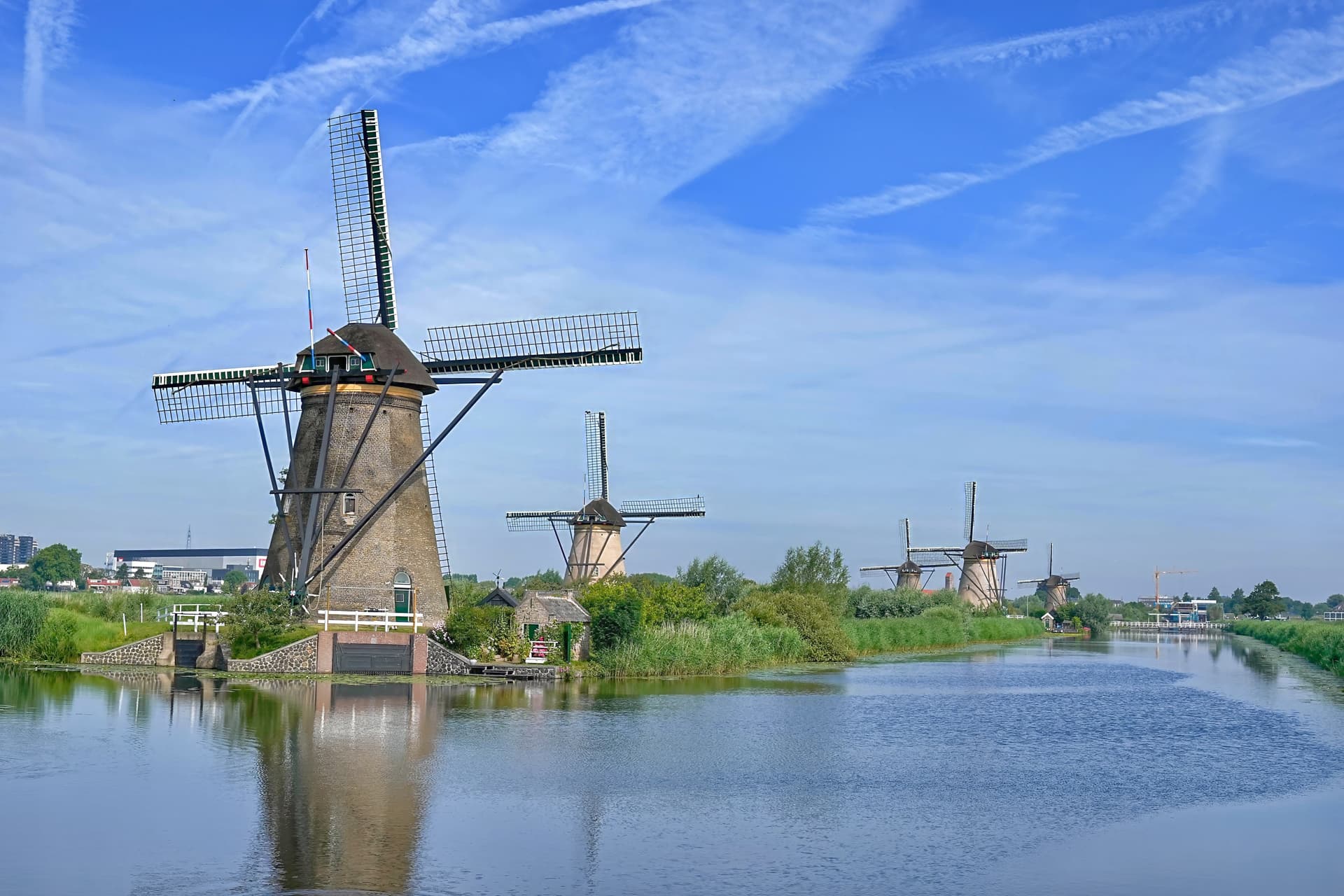 Row of historic windmills along a canal under a bright blue sky with contrails.