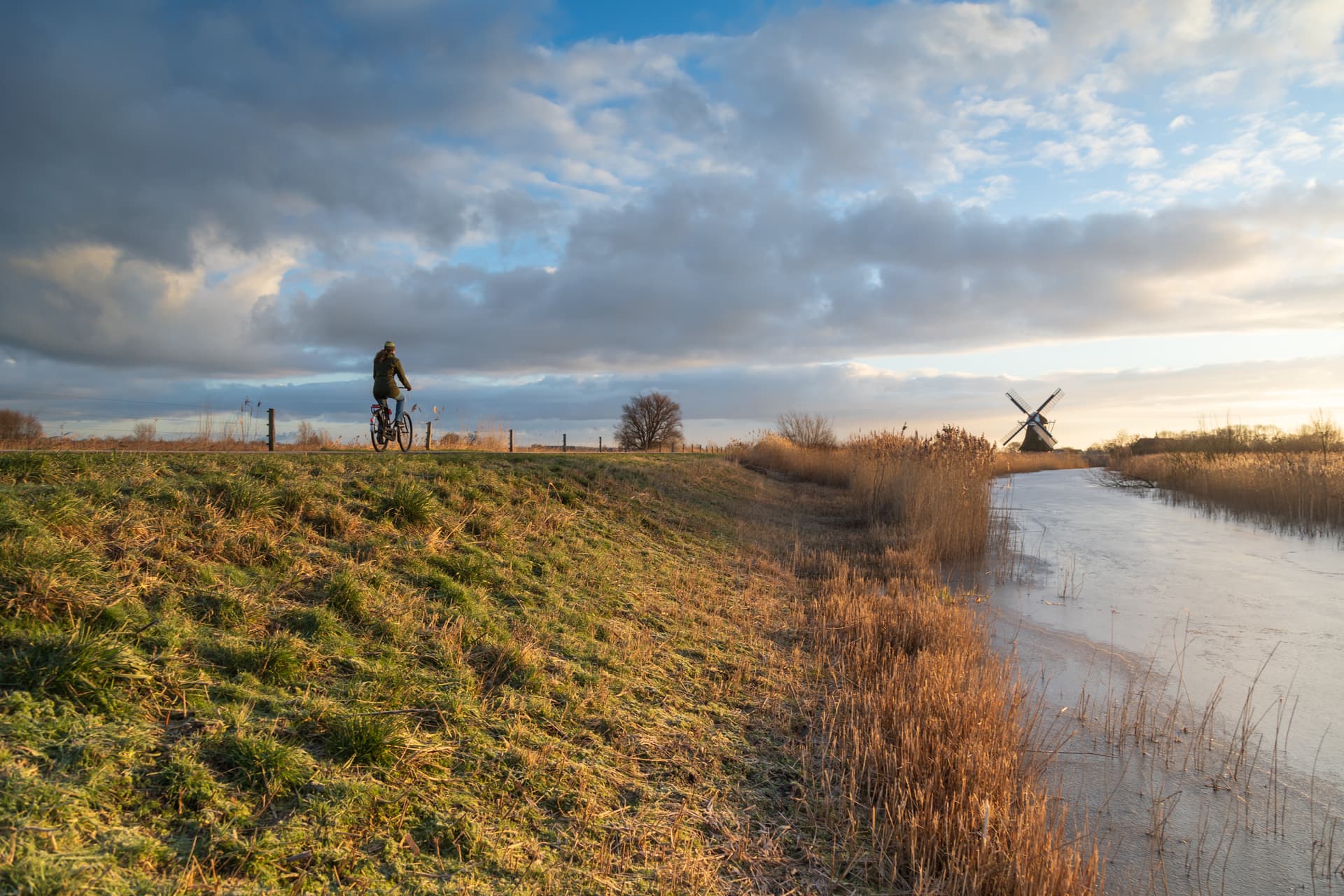 Cyclist riding past a frozen canal and windmill under dramatic winter sky