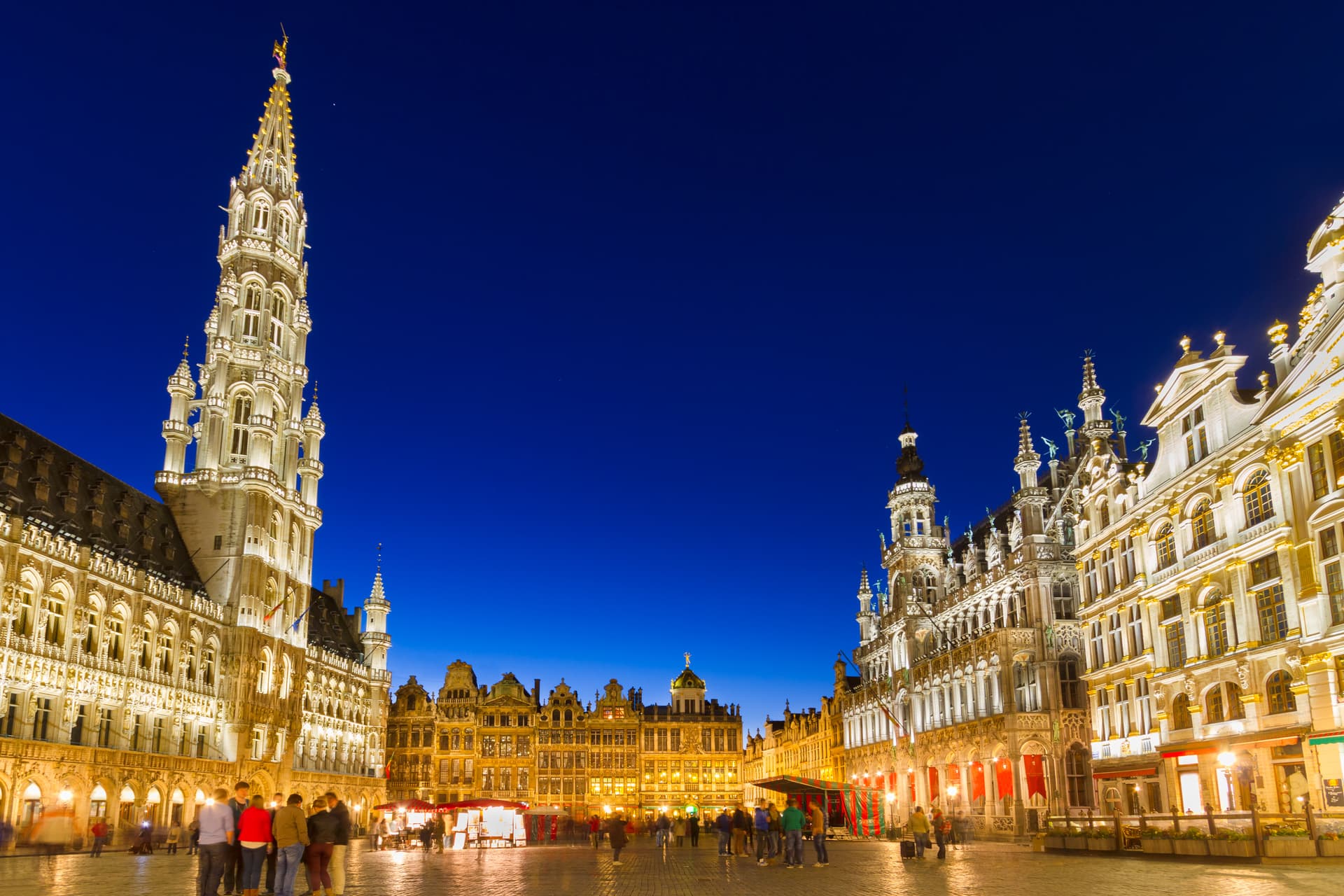 Illuminated historic buildings surrounding Brussels Grand Place at deep blue twilight with people gathered.