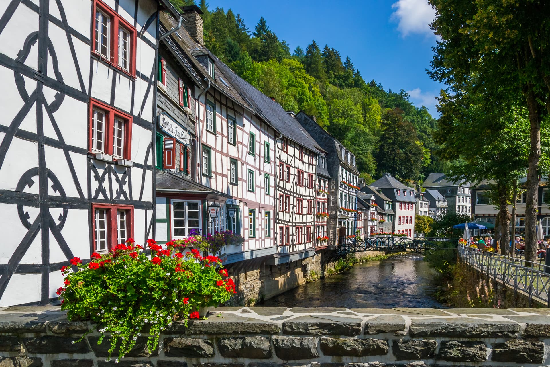 Half-timbered buildings line a river with lush green hills in the background on a sunny day.