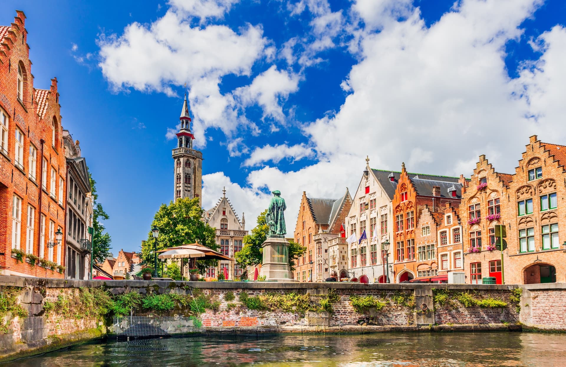 Canal view of historic brick buildings, statue, and tower under a bright blue, cloudy sky.