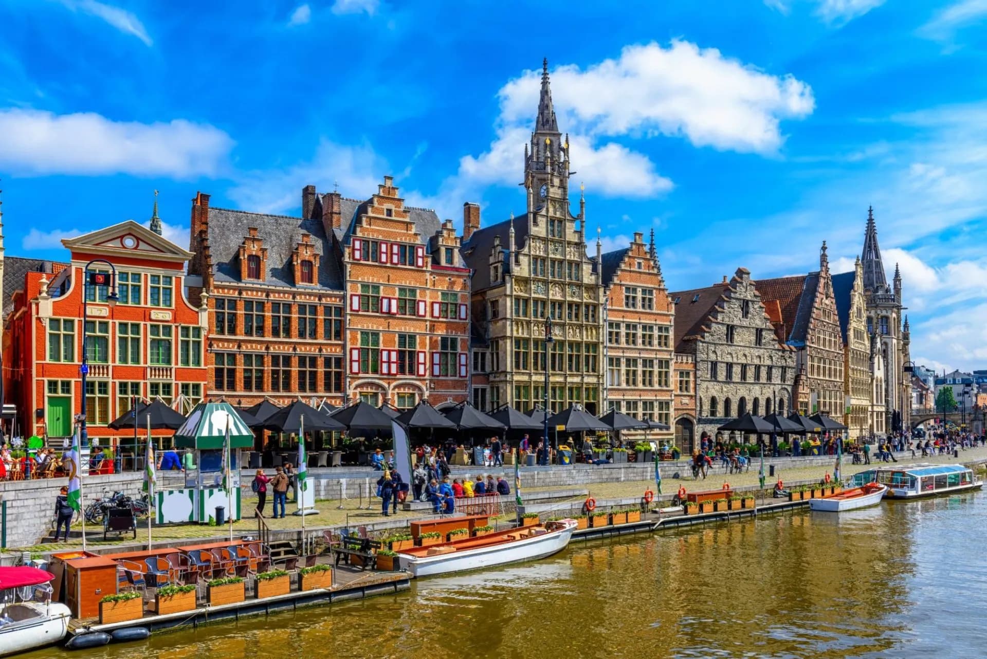 Historic guild houses line a canal in Ghent with boats docked under a bright blue, cloudy sky.