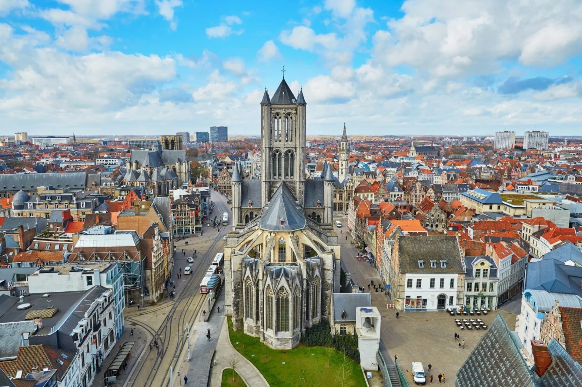 Aerial view of Ghent city center with St. Nicholas' Church tower and red rooftops under blue sky.