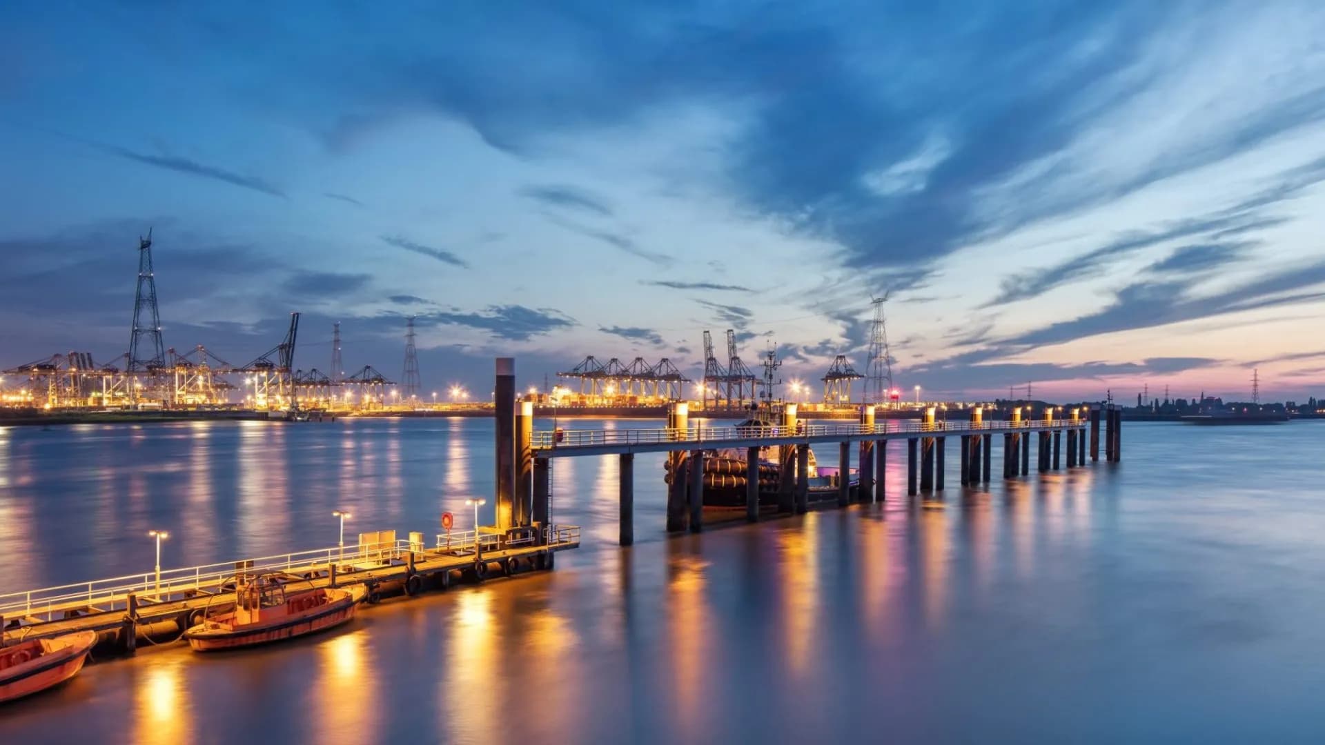 Pier with boats at dusk, illuminated industrial port with cranes across the Scheldt River.