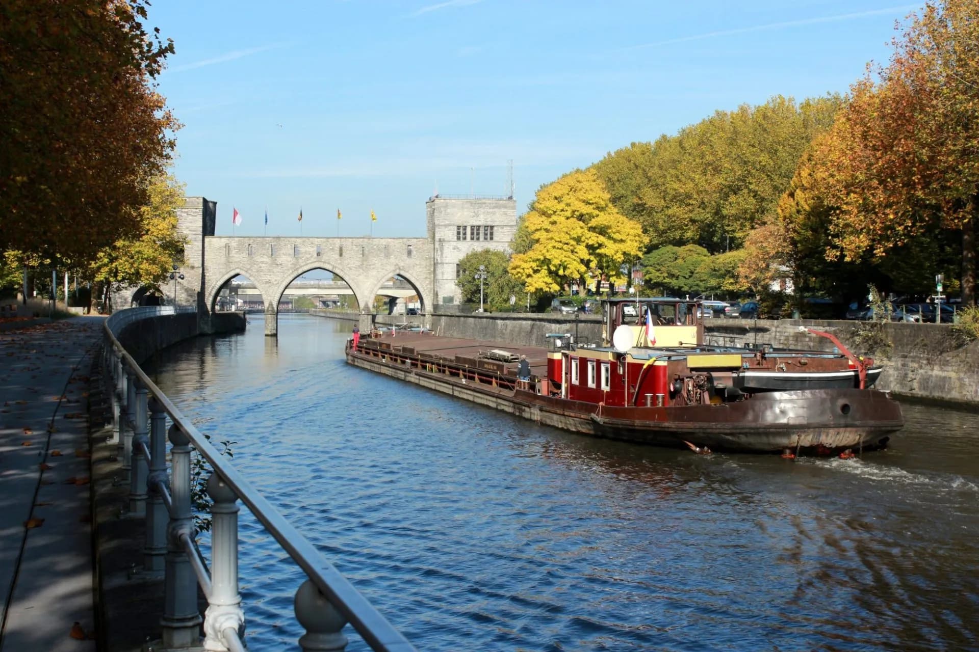 Barge moving on river past stone arch bridge with autumn trees in Tournai.