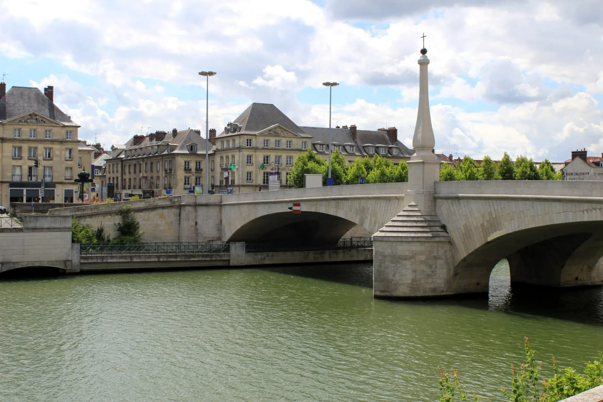 Stone bridge with cross spire over green river in Compiègne under cloudy sky