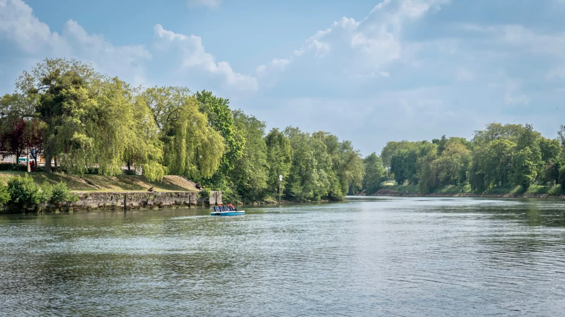 Paddle boat on river with lush green trees lining the banks under a cloudy sky.