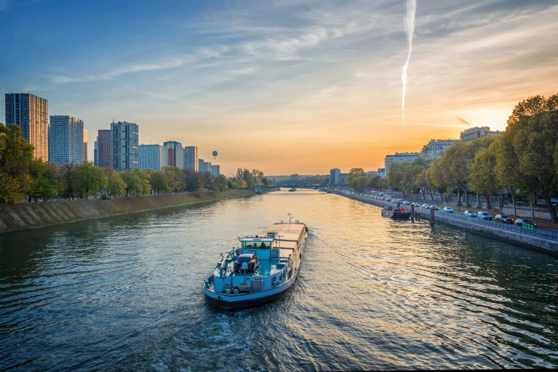Barge carrying a car travels down a river past city buildings at sunset, likely the Seine.